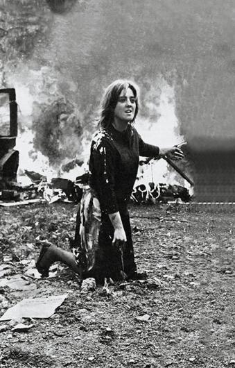 Marta Minujín, Photo from La destrucción (The Destruction), 1963. This photo is of a participatory art piece but it almost looks like a snapshot of a war scene. A woman kneels in the dirt, looking frantic while large objects burn behind her.